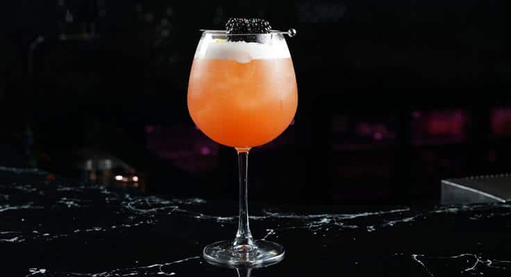 Orange cocktail with blackberry garnish in wine glass.