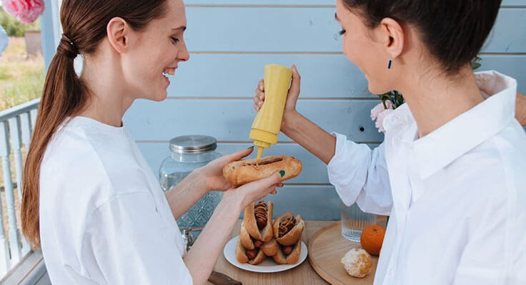 Two women preparing hot dogs with mustard.